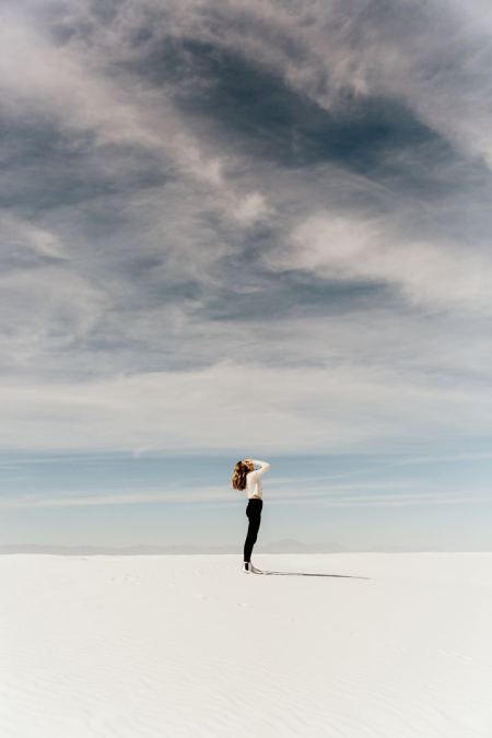 Woman on white sand looking at whisps of clouds. Photo by averie woodard on Unsplash
